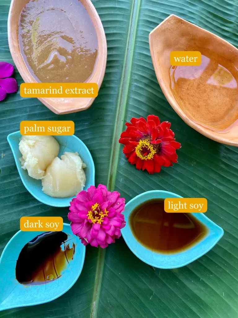 Tamarind extract and water in clay cups. Palm sugar, light soy sauce, and dark soy sauce in plastic cups. Everything is presented on a banana leaf with flowers on it.