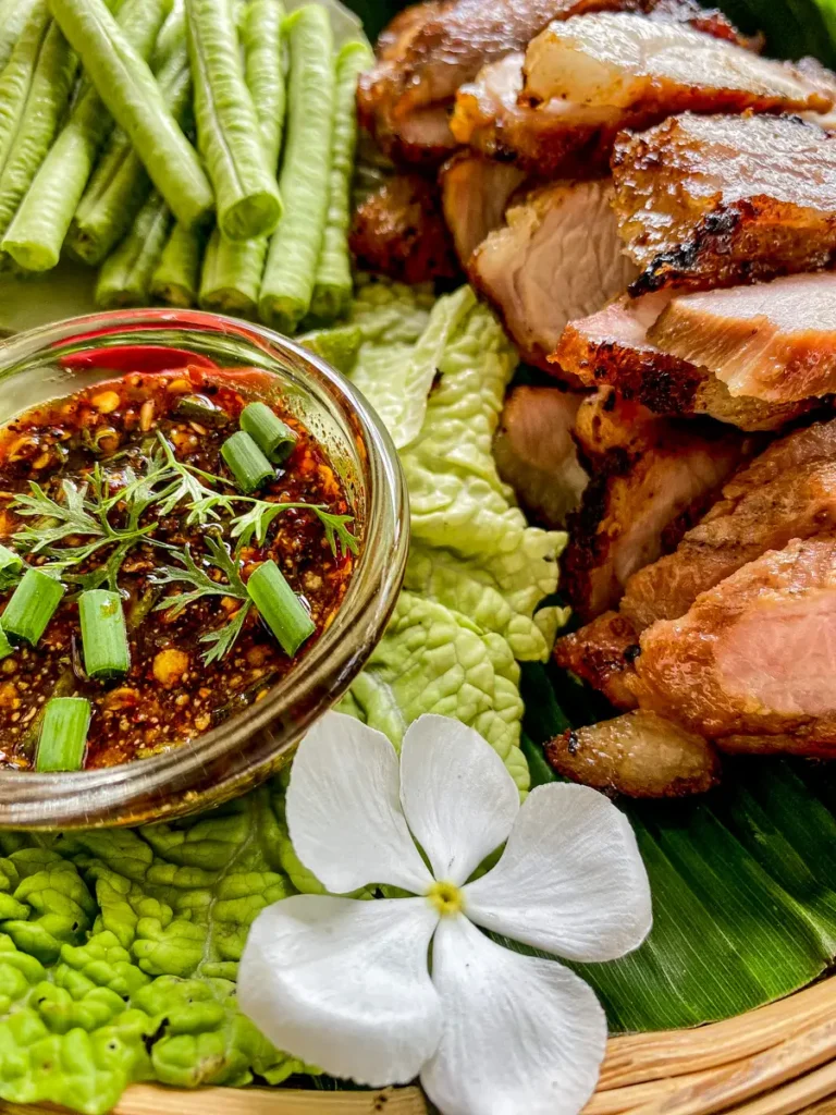 Close-up of Thai moo yang dipping sauce with chili flakes, lime juice, and herbs next to grilled pork slices and vegetables.