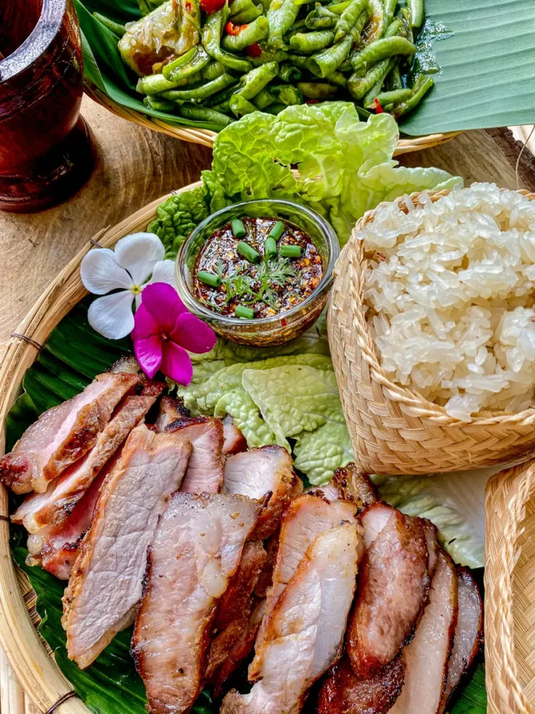 Plated Thai moo yang with sticky rice, chili dipping sauce, and long bean salad on a traditional bamboo tray.