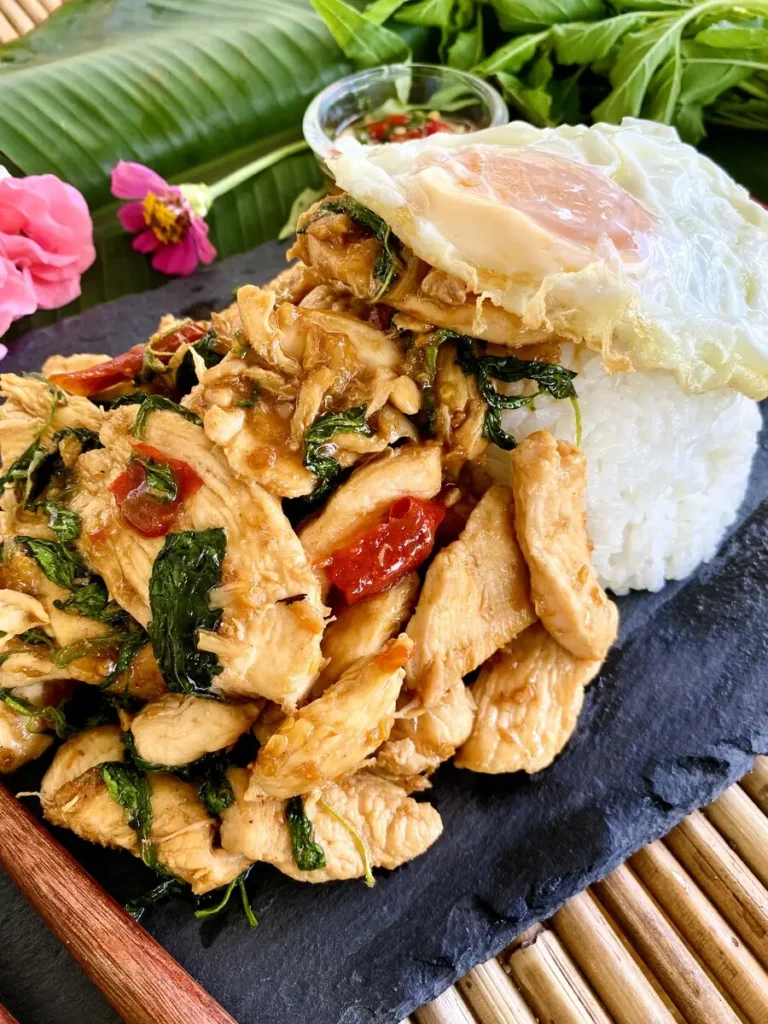 Pad krapow chicken with steamed jasmine rice and a fried egg on a black dish. The background is showing Thai holy basil leaves with flowers and prik nam pla.