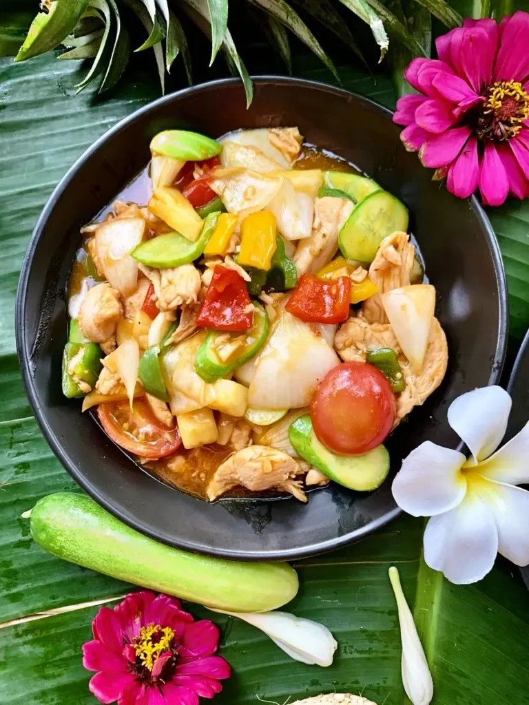 Top-down view of pad priew wan - Thai sweet and sour chicken stir-fry in a black dish with flowers and banana leaves.