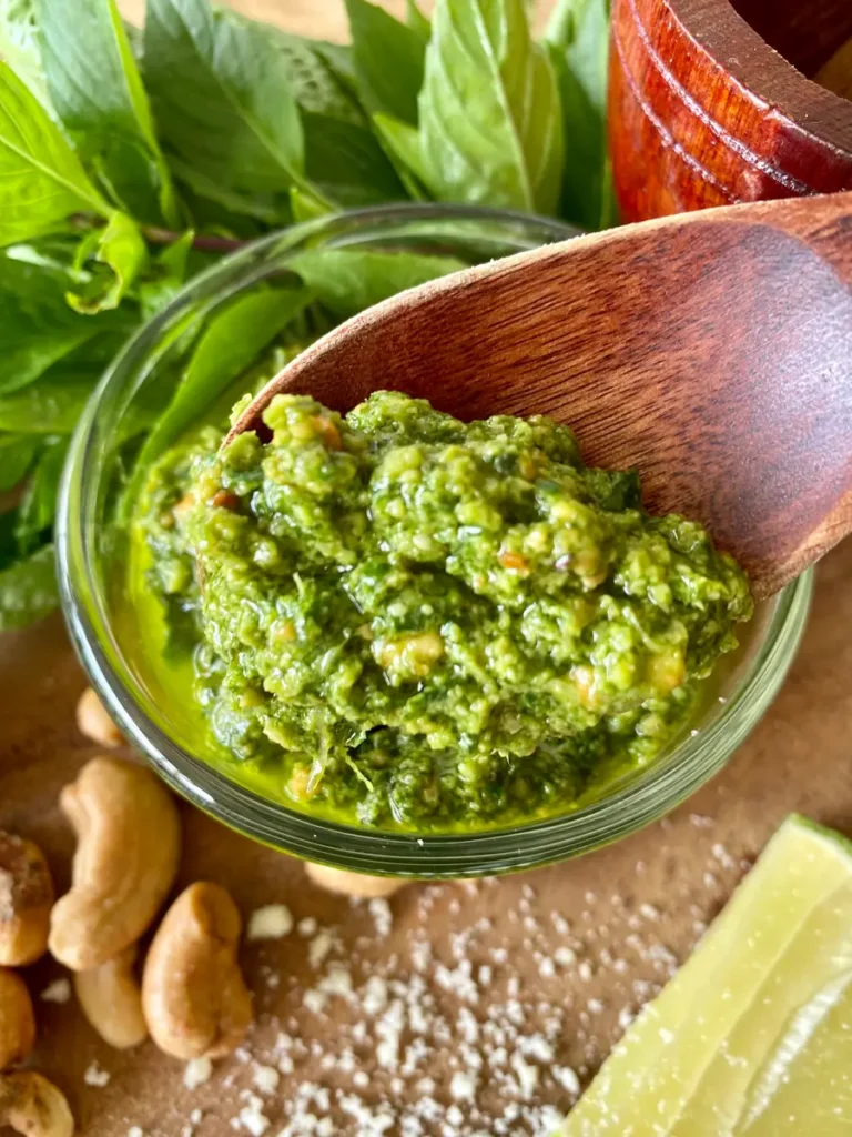 Green pesto sauce in a glass cup with a wooden spoon. Scattered around it are Thai basil leaves, peanuts, salt, and a lime wedge.