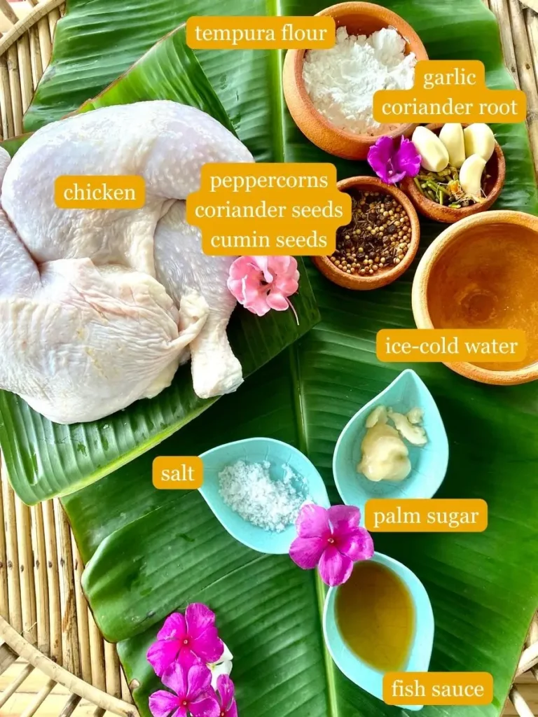 Top-down view of recipe ingredients showcased on a banana leaf; tempura flour, garlic, coriander root, peppercorns, coriander seeds, cumin seeds, chicken, water, salt, palm sugar, and fish sauce.