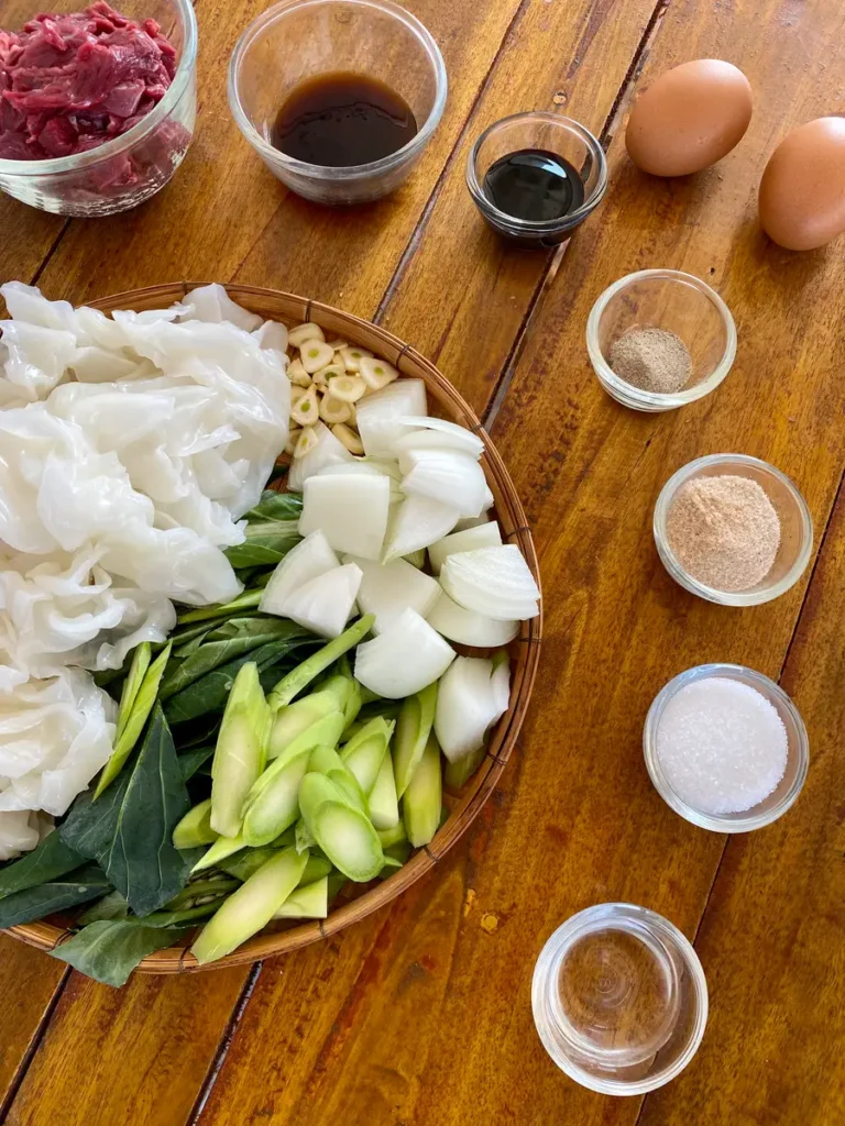 Recipe ingredients on a wooden table; 2 eggs, sliced beef, dark soy, oyster sauce, white pepper, flavor seasoning, salt, onion, garlic, white sugar, Chinese broccoli, and fresh rice noodles.