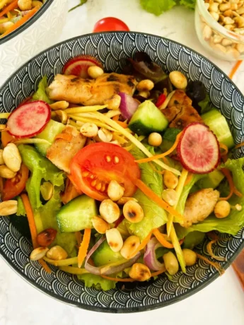Spicy Thai chicken salad with honey, tomatoes, peanuts, cucumbers, radish, cabbage, and more vegetables in a blue and white bowl. In the top are roasted peanuts, another bowl, and some fresh vegetables.