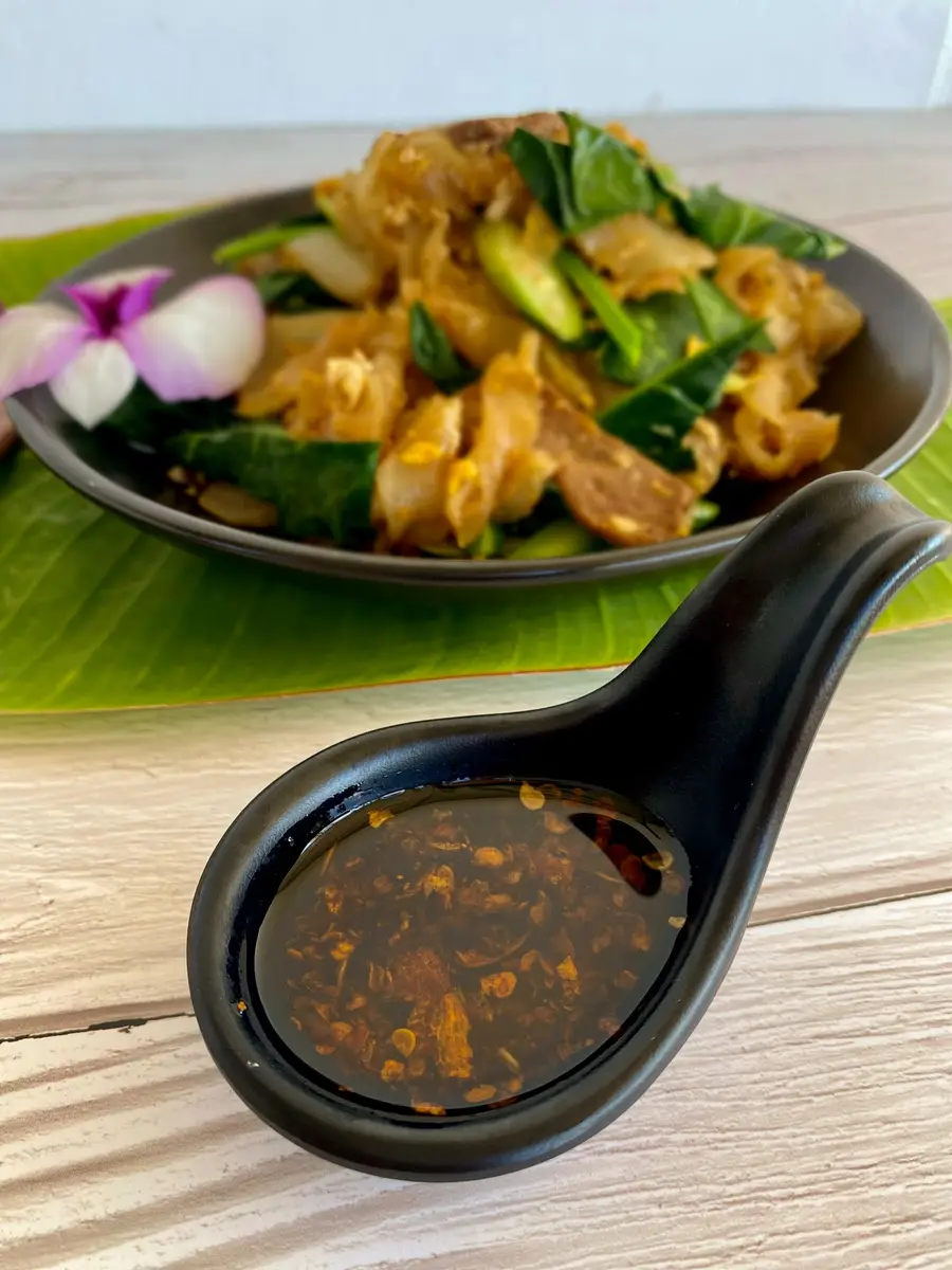 Large black spoon containing oil with red chili flakes. In the distance is a black dish with stir-fried noodles and vegetables.