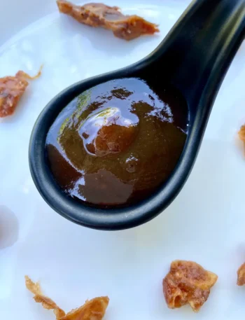 Thick and glossy homemade tamarind sauce in a black ceramic spoon, with tamarind pods scattered on a white background.