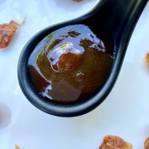 Thick and glossy homemade tamarind sauce in a black ceramic spoon, with tamarind pods scattered on a white background.