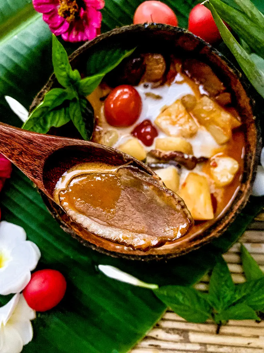 Thai duck curry with coconut milk and pineapple, featuring a wooden spoon lifting a slice of roasted duck from the curry bowl.
