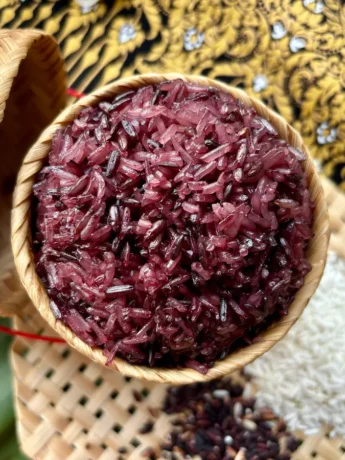 Authentic Thai purple sticky rice in a bamboo serving basket, with raw white and black rice grains spread out on a bamboo mat next to it.