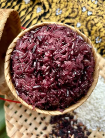 Authentic Thai purple sticky rice in a bamboo serving basket, with raw white and black rice grains spread out on a bamboo mat next to it.