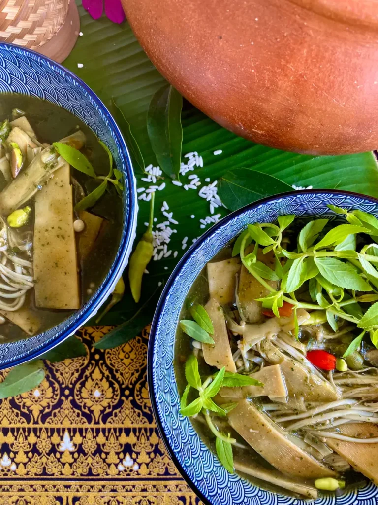 2 Portions of Thai bamboo soup served in bowls, garnished with vegetables. Scattered around it are fresh chilies, glutinous rice, and more.