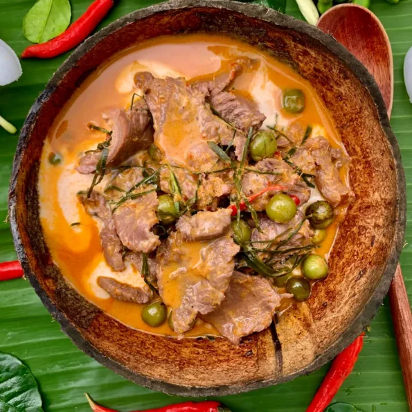 Top-down view of Thai beef panang curry, garnished with red chilies and kaffir lime leaf strips, in a coconut shell. Alongside it are rice vermicelli noodles and a wooden spoon.
