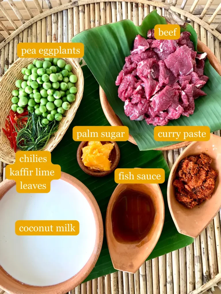 Bird's eye view of beef panang curry ingredients; beef, pea eggplants, palm sugar, curry paste, chilies, kaffir lime leaves, fish sauce, and coconut milk.
