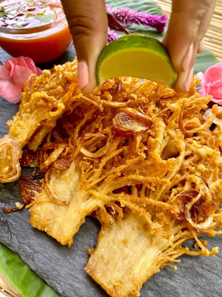 Several deep-fried enoki mushrooms arranged on a black dish, with a hand squeezing fresh lime juice on top.