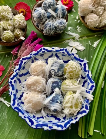 Khanom tom - Thai coconut balls served in a blue and white bowl on a banana leaf. In the distance are more colorful coconut balls, served in coconut shells.