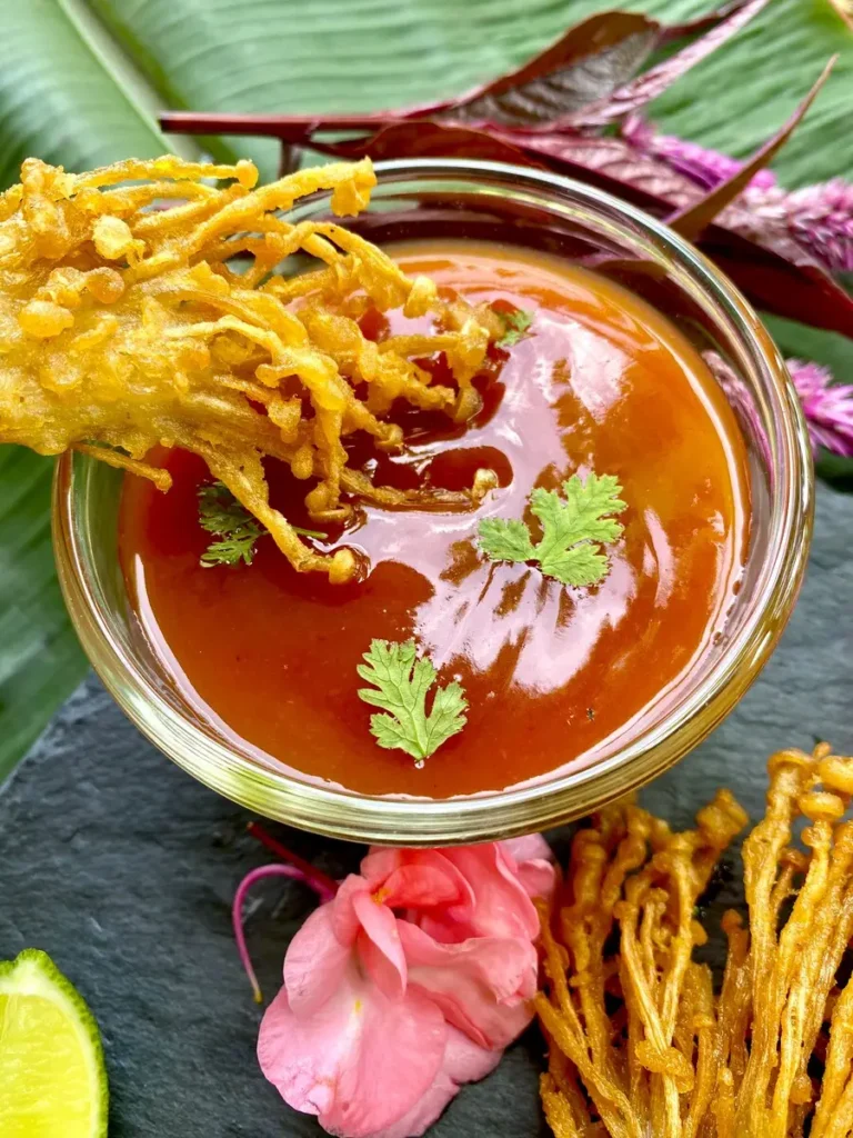 Fried enoki mushrooms dipped into a glass cup with spicy chili sauce.