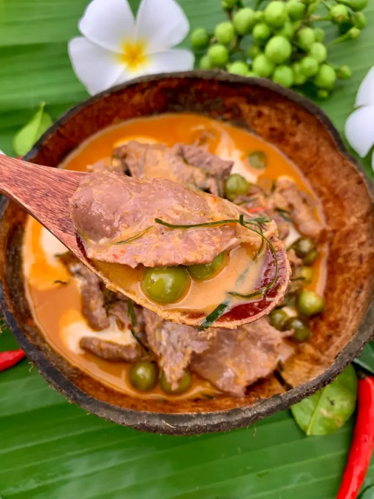 Panang beef curry in a coconut shell with a wooden spoon, lifting a spoonful of the dish.