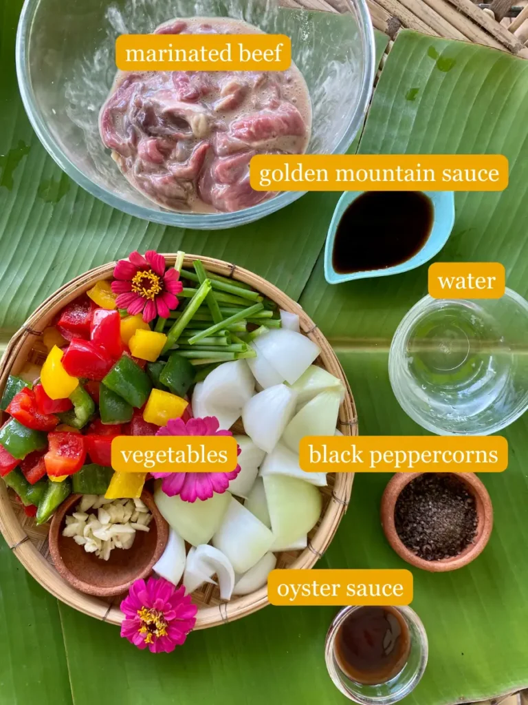 Bird's eye view of recipe ingredients displayed on a banana leaf; marinated beef, golden mountain sauce, water, vegetables, black peppercorns, and oyster sauce. 