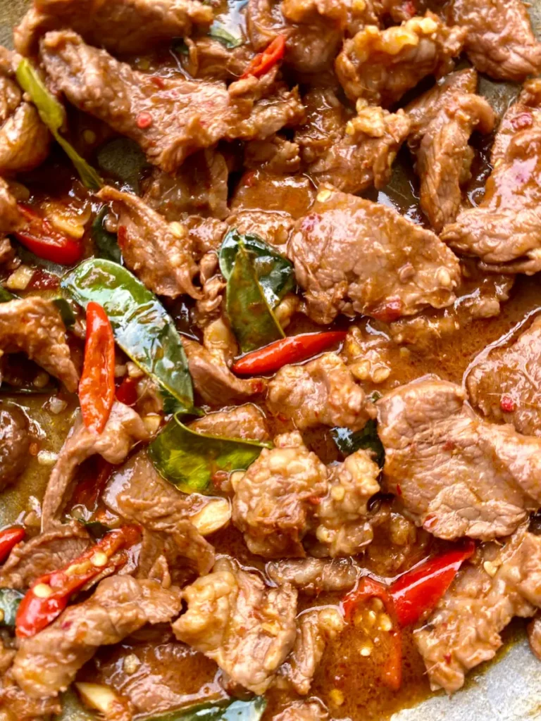 Close-up of spicy Thai beef slices coated with a dark sauce, kaffir lime leaves, and red chilies.
