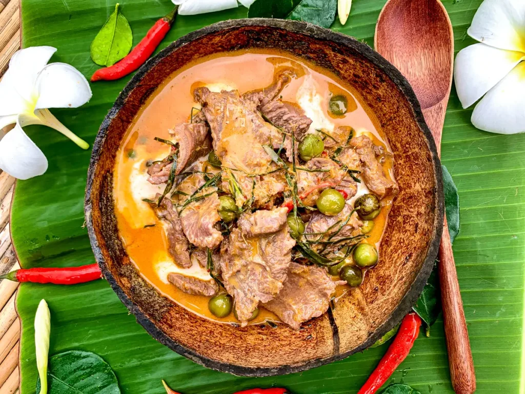 Thai beef panang curry with rich coconut milk, green pea eggplants, and red curry sauce on a banana leaf background.