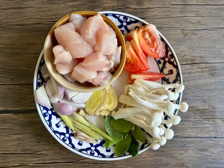 Prepped ingredients on a blue and white Thai plate: chicken, tomato, shallots, galangal, lemongrass, mushrooms, and lime leaves.