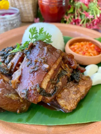 Thai braised pork belly dish served on a banana leaf with sticky rice, fresh cilantro, and a side of spicy vinegar dipping sauce.