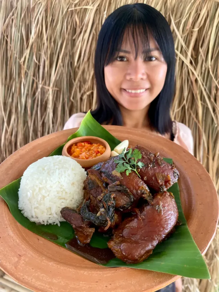 Smiling woman presenting a plate of Thai braised pork belly with rice on a banana leaf, complete with dipping sauce.