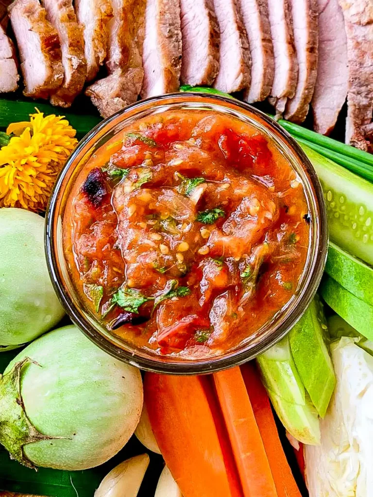Jeow mak len chili dip in a glass bowl with grilled pork and raw vegetables.