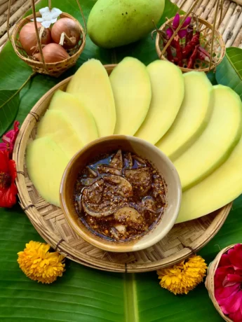 Sliced mango arranged around jeow mak muang, presented on a woven bamboo tray with onions and dried chilies.