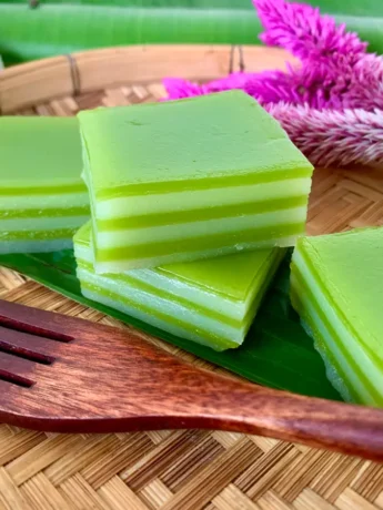 Green khanom chan, Thai layered dessert, stacked with tropical flowers and greenery in the background.