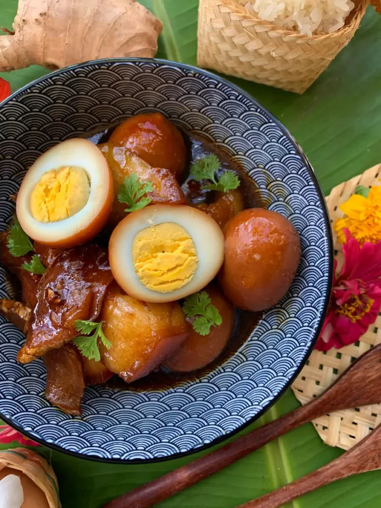 Close-up view of glossy braised pork belly pieces and golden halved hard-boiled eggs in a patterned bowl, accompanied by ginger and sticky rice.