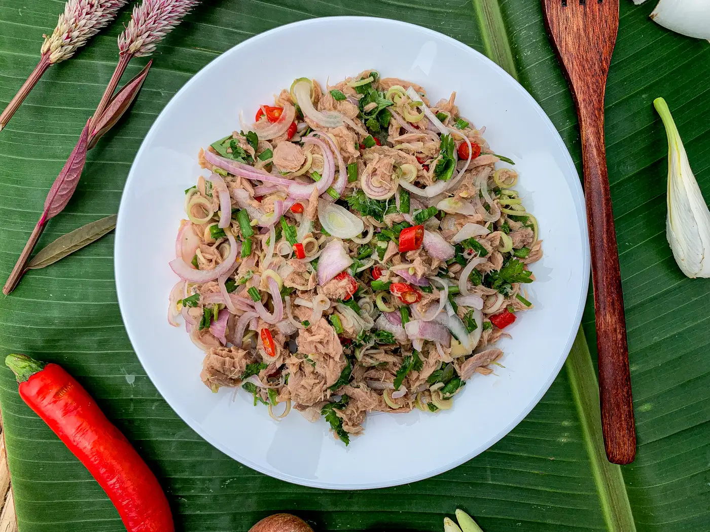 Thai tuna salad made with herbs, shallots, and chili on a white plate surrounded by vegetables.