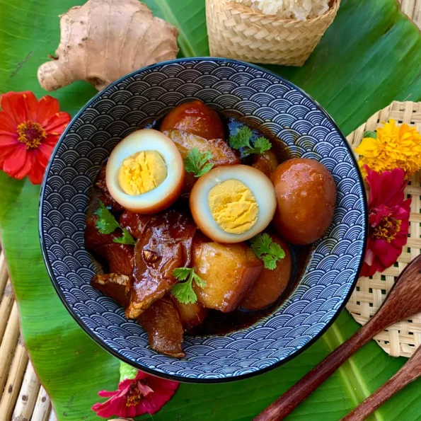 Overhead shot of tom khem, a Lao braised pork with egg dish, served with sticky rice, surrounded by fresh ingredients, all set on a banana leaf.