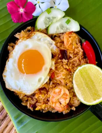 Tom yum fried rice served in a black bowl with a sunny-side-up egg, fresh shrimp, and cucumber slices, garnished with flowers.