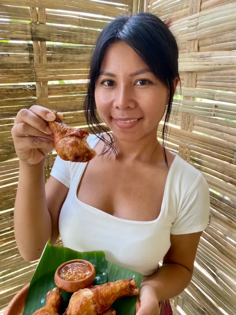 Thai woman holding a crispy fried chicken drumsticks with dipping sauce, against a bamboo background.