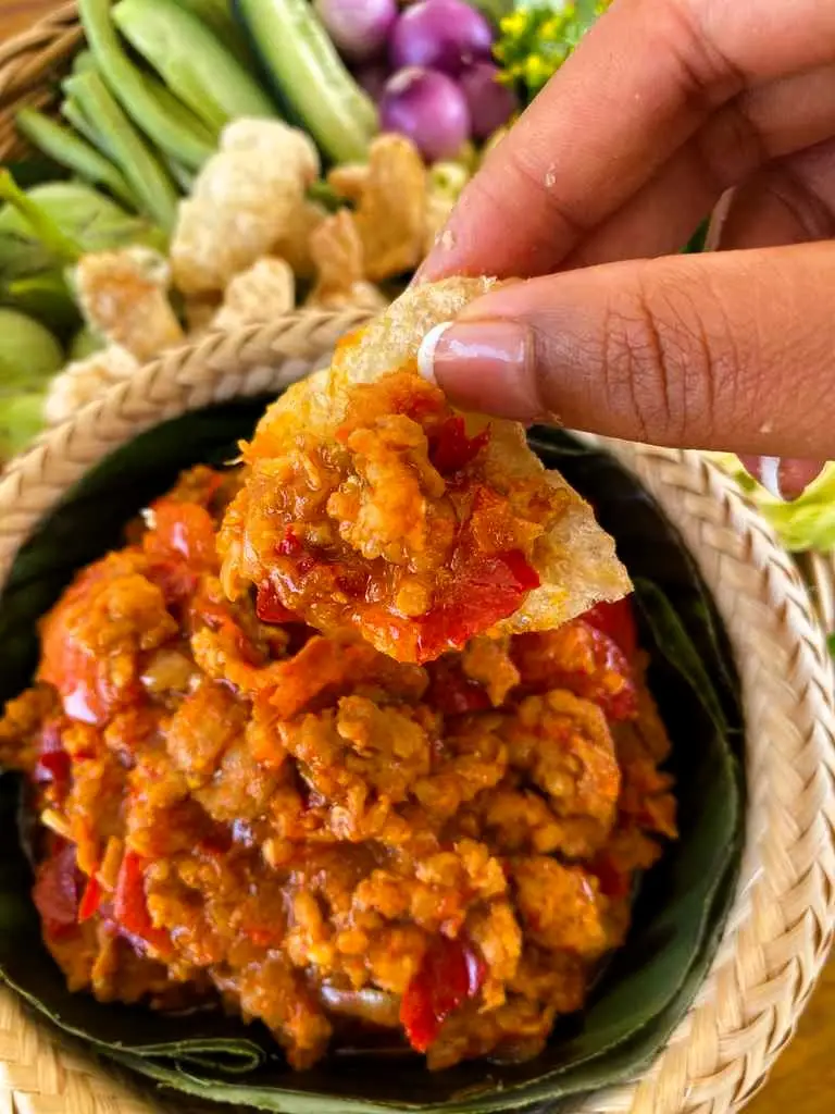 Hand dipping a pork crackling into nam prik ong, a Northern Thai dipping sauce with minced pork and tomatoes.