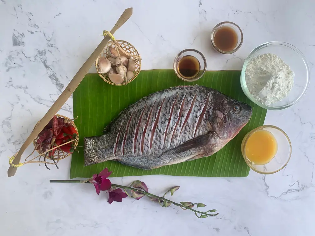 Ingredients for Thai spicy fish recipe arranged on a marble counter, featuring a whole fish with tamarind sauce, rice flour, garlic, dried chilies, and more.