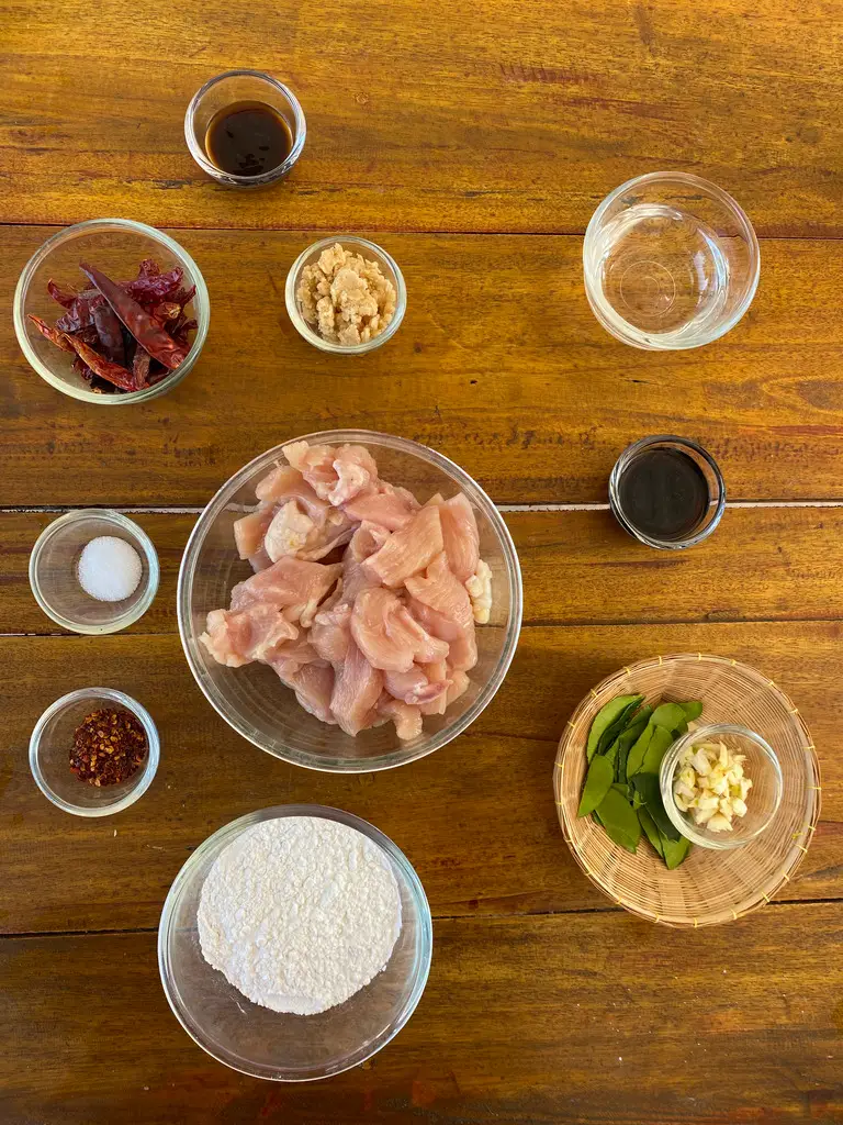 Top view of Thai recipe ingredients on a wooden table: soy sauce, dried chilies, sugar, chicken, water, salt, crispy flour, chilies, garlic, and kaffir lime leaves.