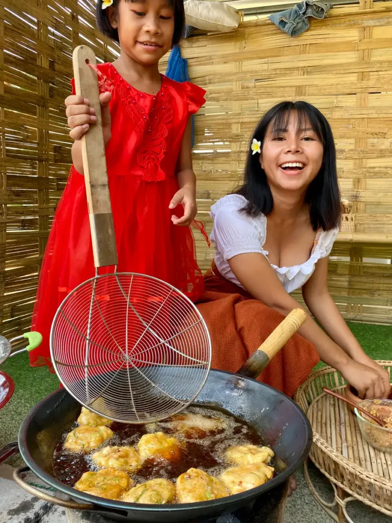 A Thai woman and a young girl smiling while cooking tod mun pla, Thai fish cakes, in a large wok outdoors.