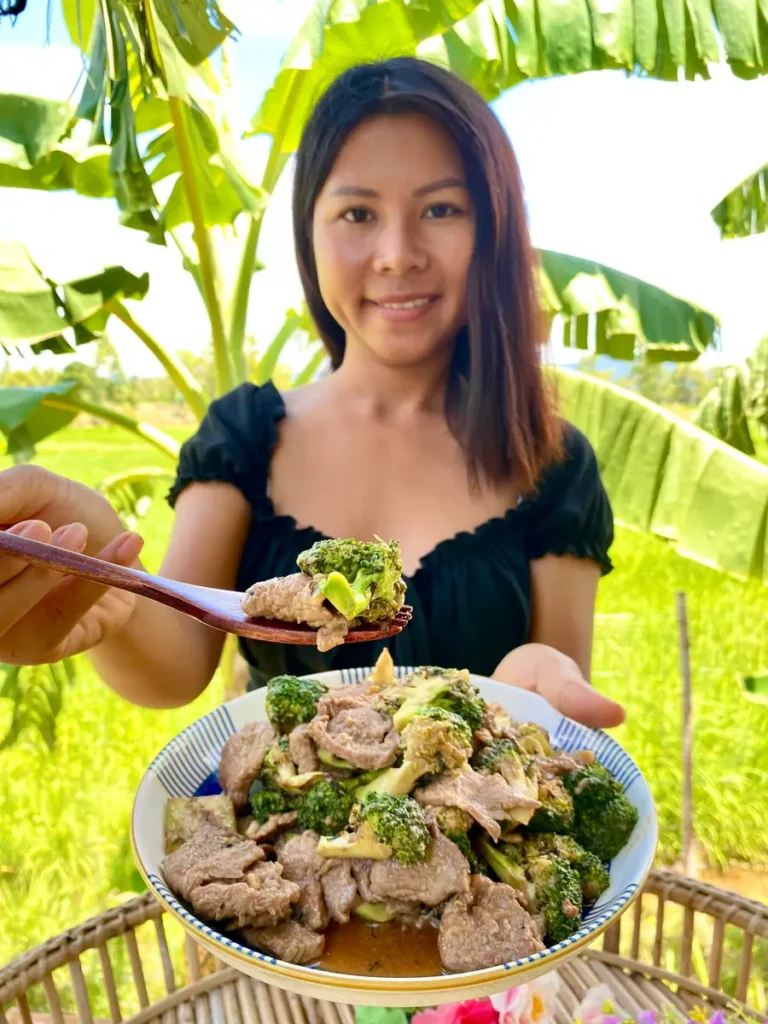 Thai woman smilingly presenting a white dish with stir-fried broccoli and beef, with a rural background.