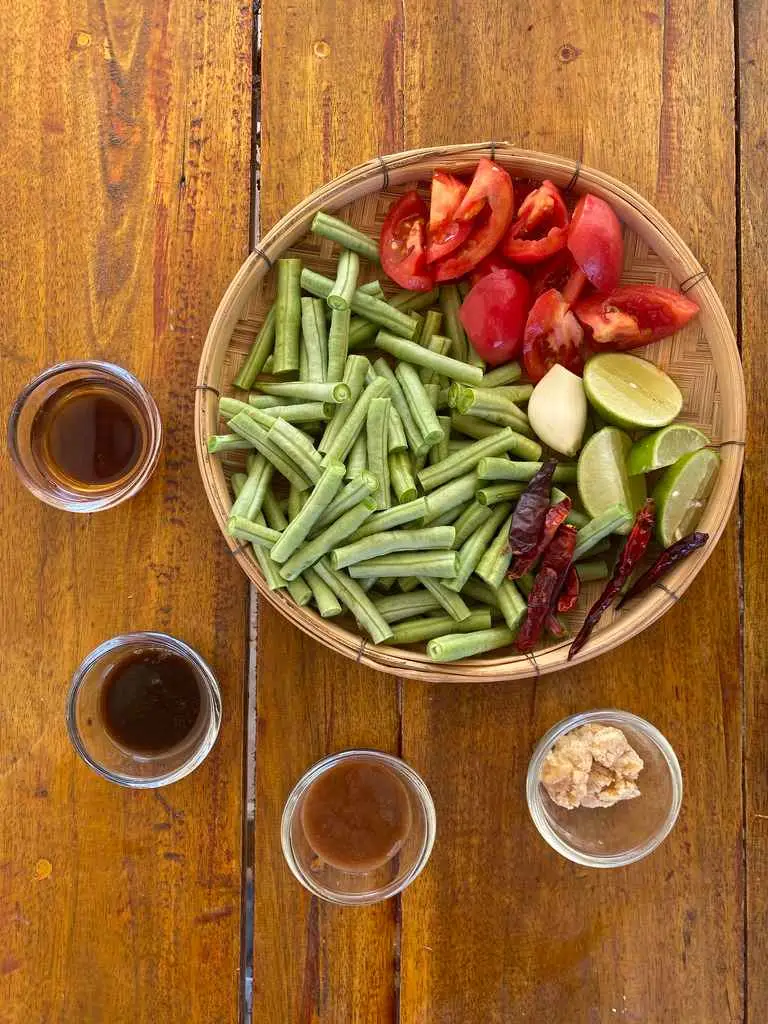 Top-view of ingredients for Thai long bean salad: yardlong beans, lime, tomatoes, dried chilies, fish sauce, palm sugar, tamarind sauce, and more.