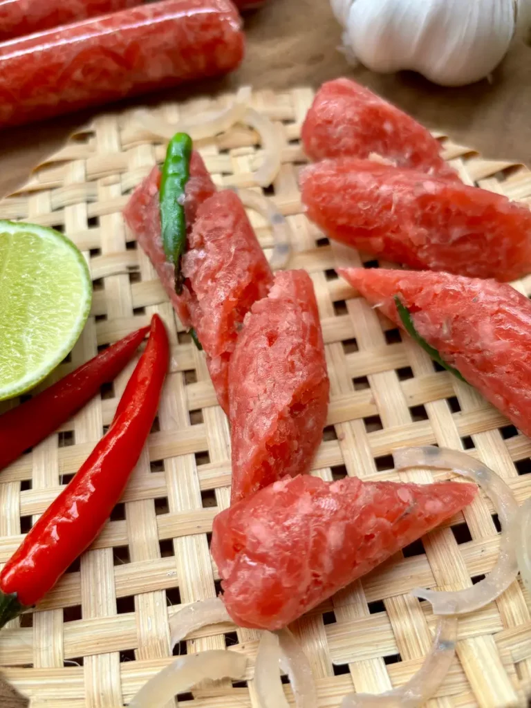 Close-up of Lao som moo sausage slices with chilies, lime wedge, and fresh garlic, ready for a Thai meal.