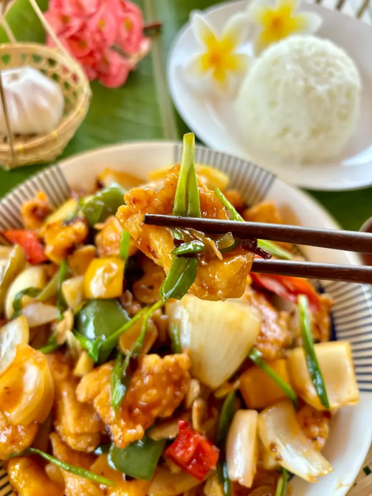 Close-up of Thai chili paste stir-fry with a focus on a piece of chicken picked up with chopsticks, alongside jasmine rice and Thai decor in the background.