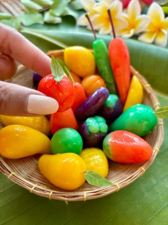 Colorful Thai luk chup mung bean dessert shaped like fruits and vegetables in a woven basket on a banana leaf background.