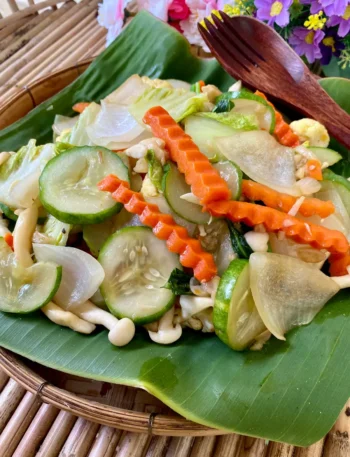 Thai vegetables stir-fry, pad pak ruam mit, served on a banana leaf with a wooden fork, showcasing carrots, cucumbers, onions, and mushrooms.