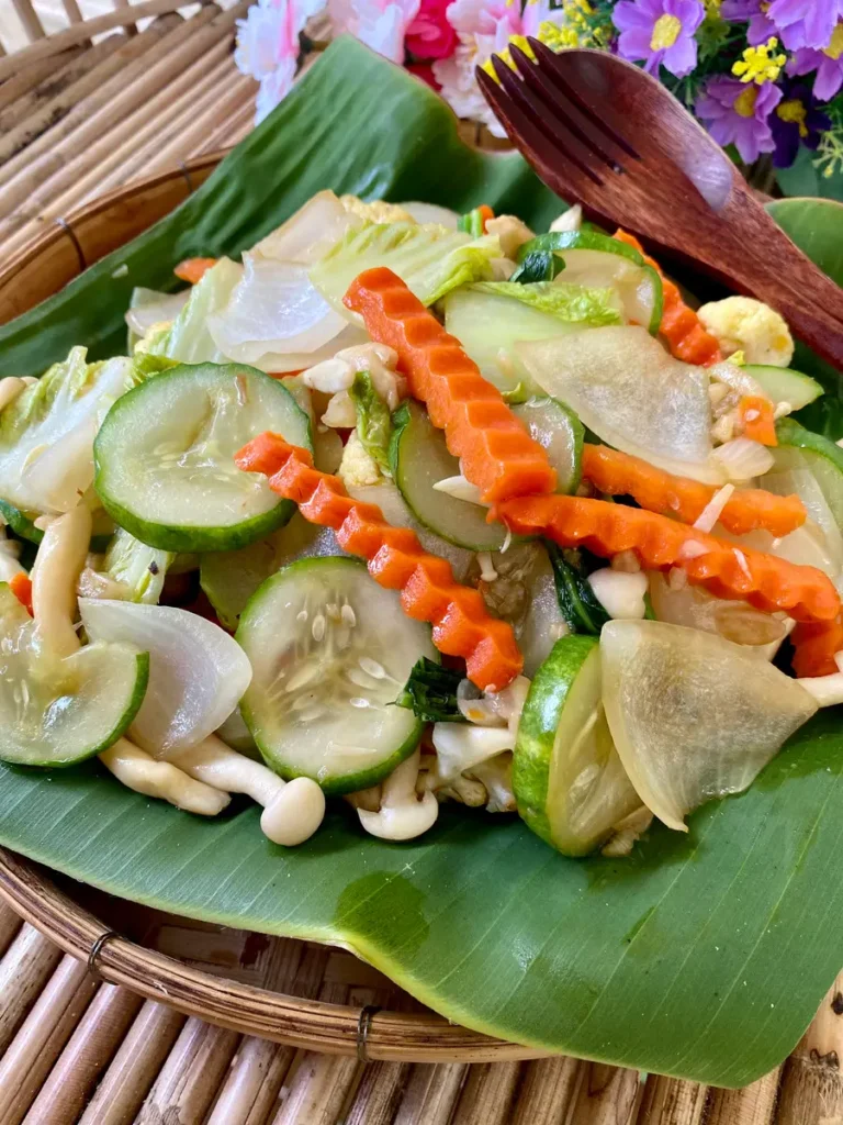 Thai vegetables stir-fry, pad pak ruam mit, served on a banana leaf with a wooden fork, showcasing carrots, cucumbers, onions, and mushrooms.