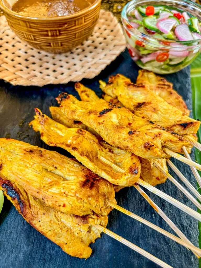 Chicken satay skewers with creamy peanut sauce and fresh cucumber salad on a black slate plate.