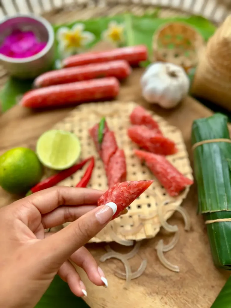 A hand holding a slice of Lao som moo fermented pork sausage with fresh ingredients in the background.