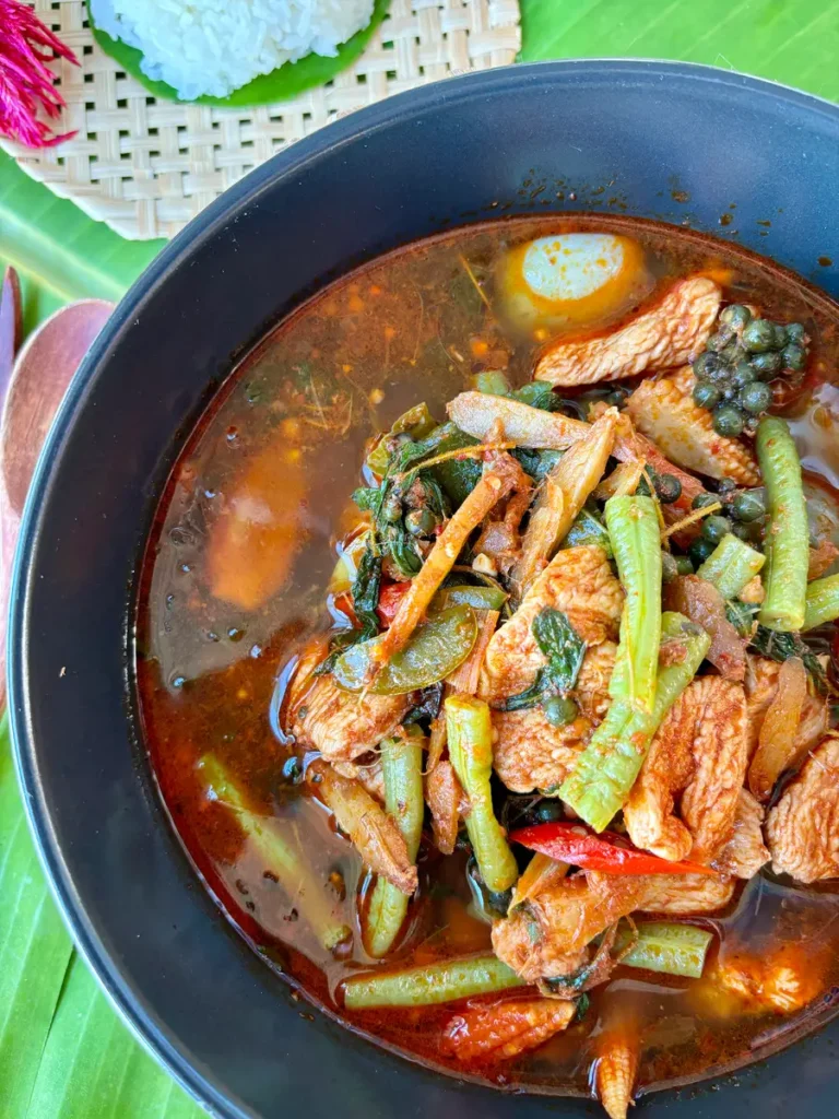 Gaeng pa curry served in a large black bowl, with a spicy broth, yard long beans, chicken, chilies, fresh peppercorns, fingerroot, and more spices.